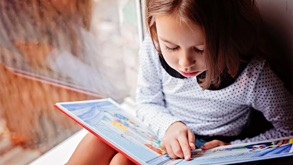 Toddler girl learning how to read a book pointing and sounding out the words.